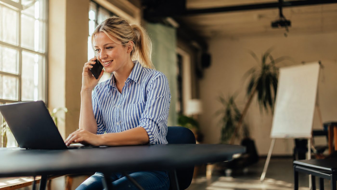 A woman at a laptop in an office, smiling while talking on her phone.